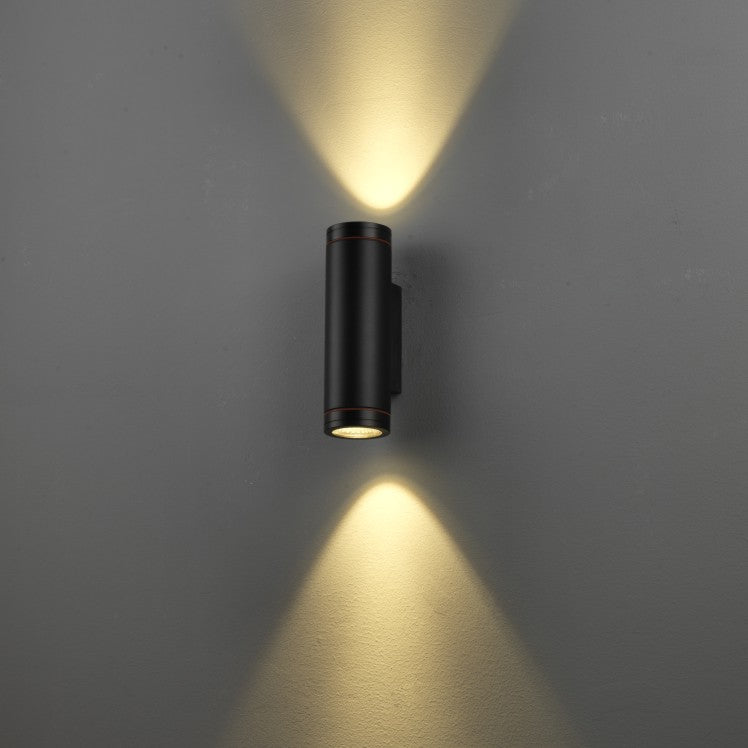 A modern black cylindrical wall sconce light fixture mounted on a gray wall, emitting warm yellow light upwards and downwards in a V-shape pattern.