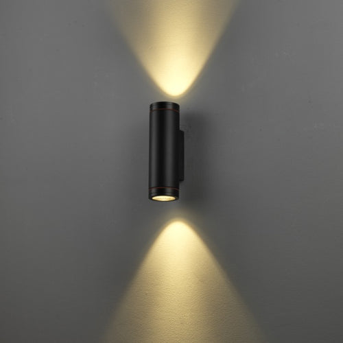 A modern black cylindrical wall sconce light fixture mounted on a gray wall, emitting warm yellow light upwards and downwards in a V-shape pattern.