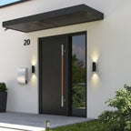 Modern black front door with a vertical copper-toned handle, set in a white stucco entryway with wall-mounted lights, a mailbox, and the house number 20.