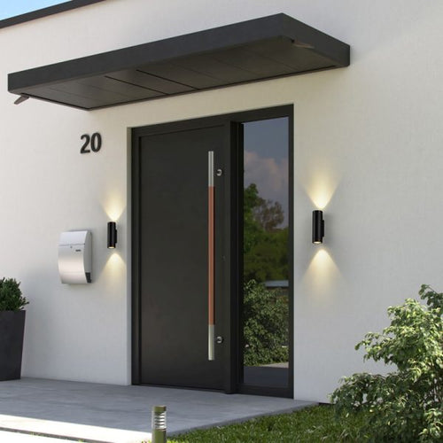 Modern black front door with a vertical copper-toned handle, set in a white stucco entryway with wall-mounted lights, a mailbox, and the house number 20.